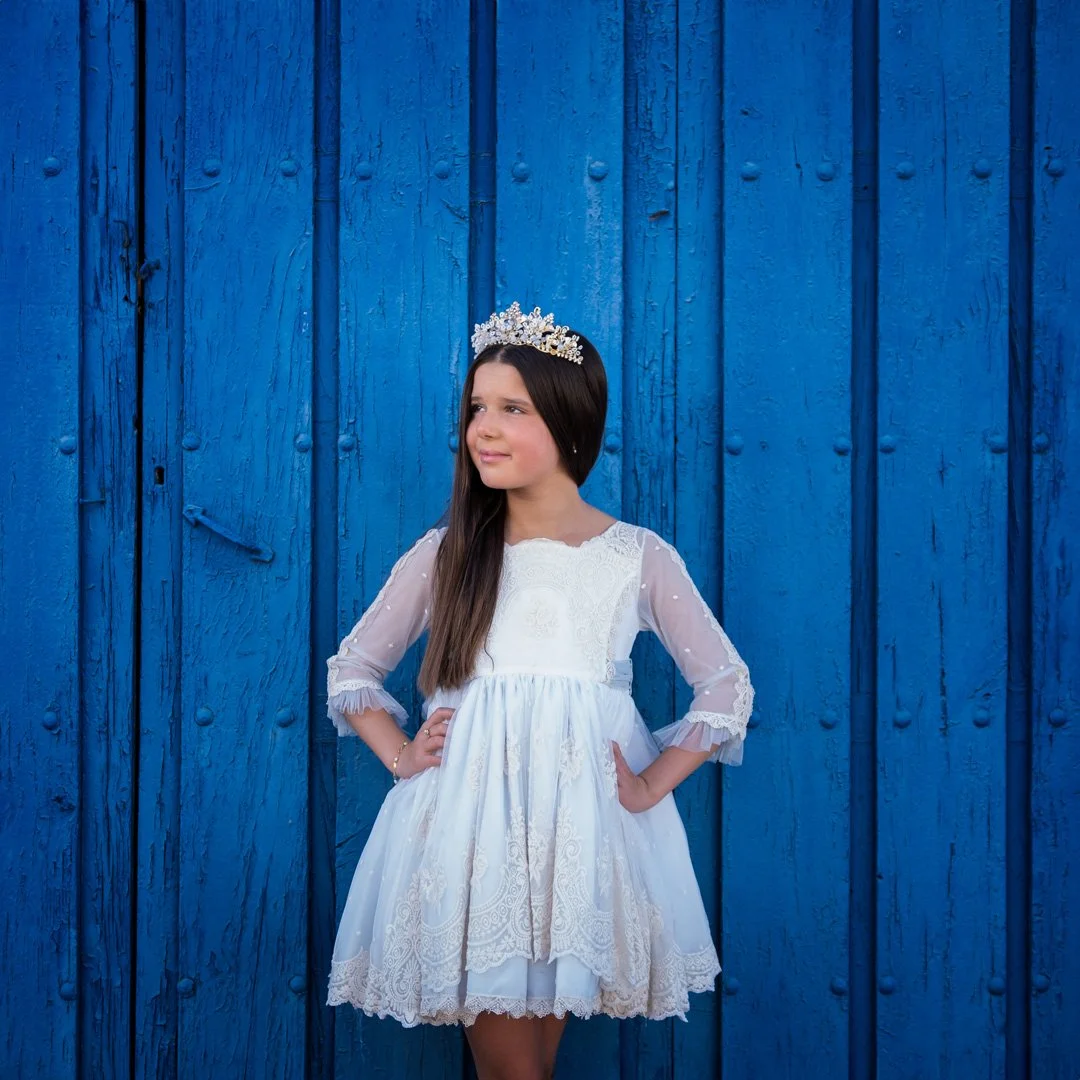 Niña con vestido blanco, diadema y manos en las caderas, de pie frente a una cerca de madera azul.