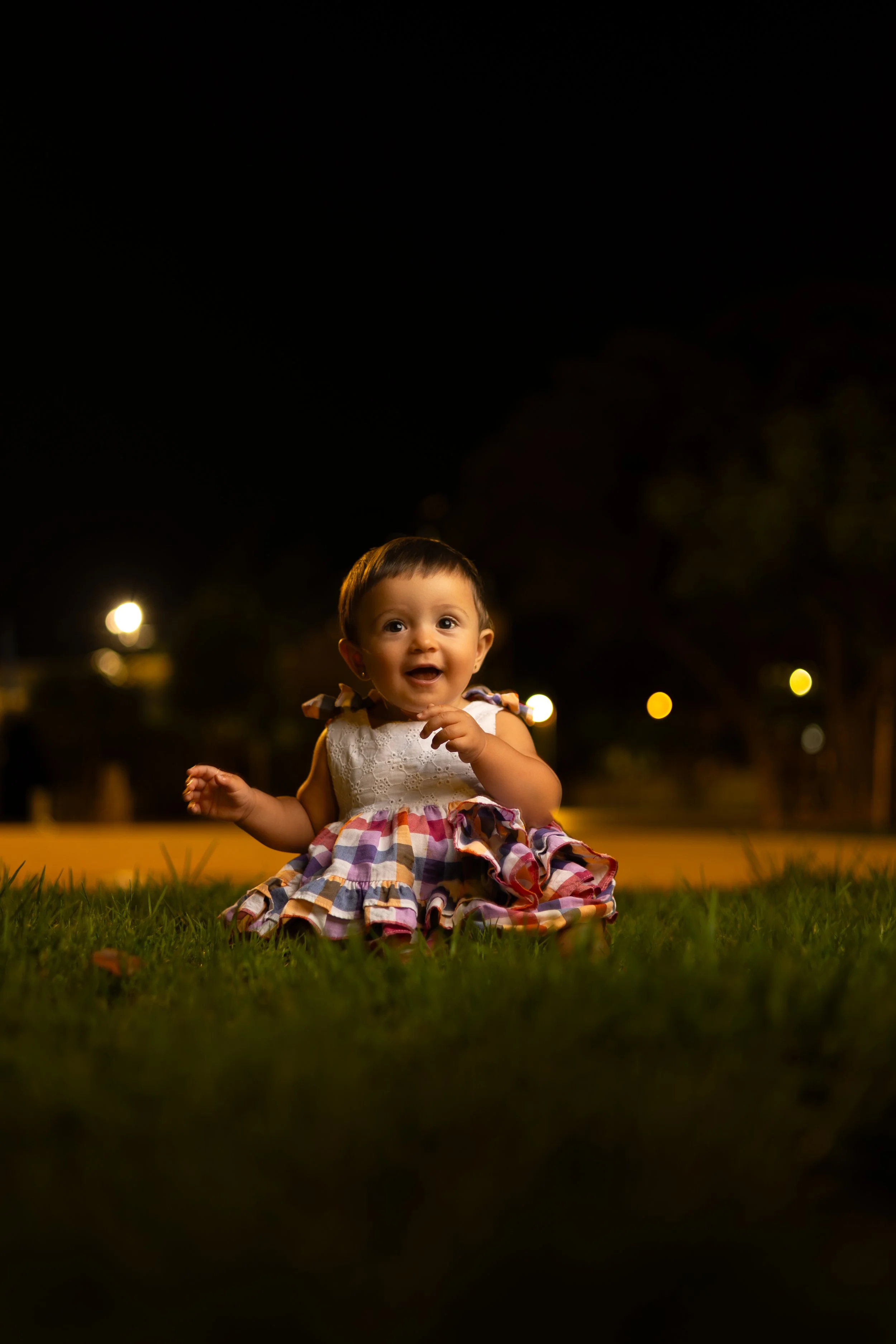 Bebé sonriendo y gateando en un parque durante la noche, con fondo oscuro y luces difusas.