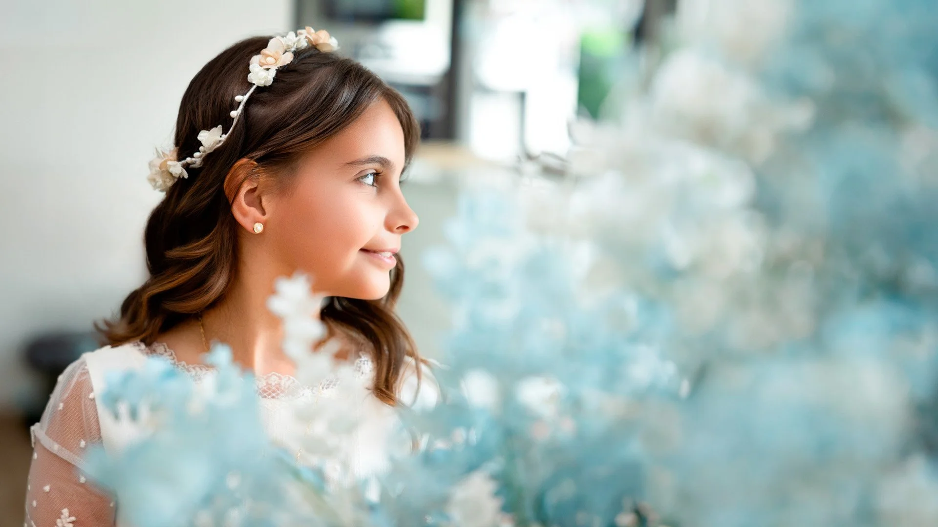 Joven mujer con rulos sueltos, usando una diadema con flores y pendientes de perla, sonriendo delicadamente.