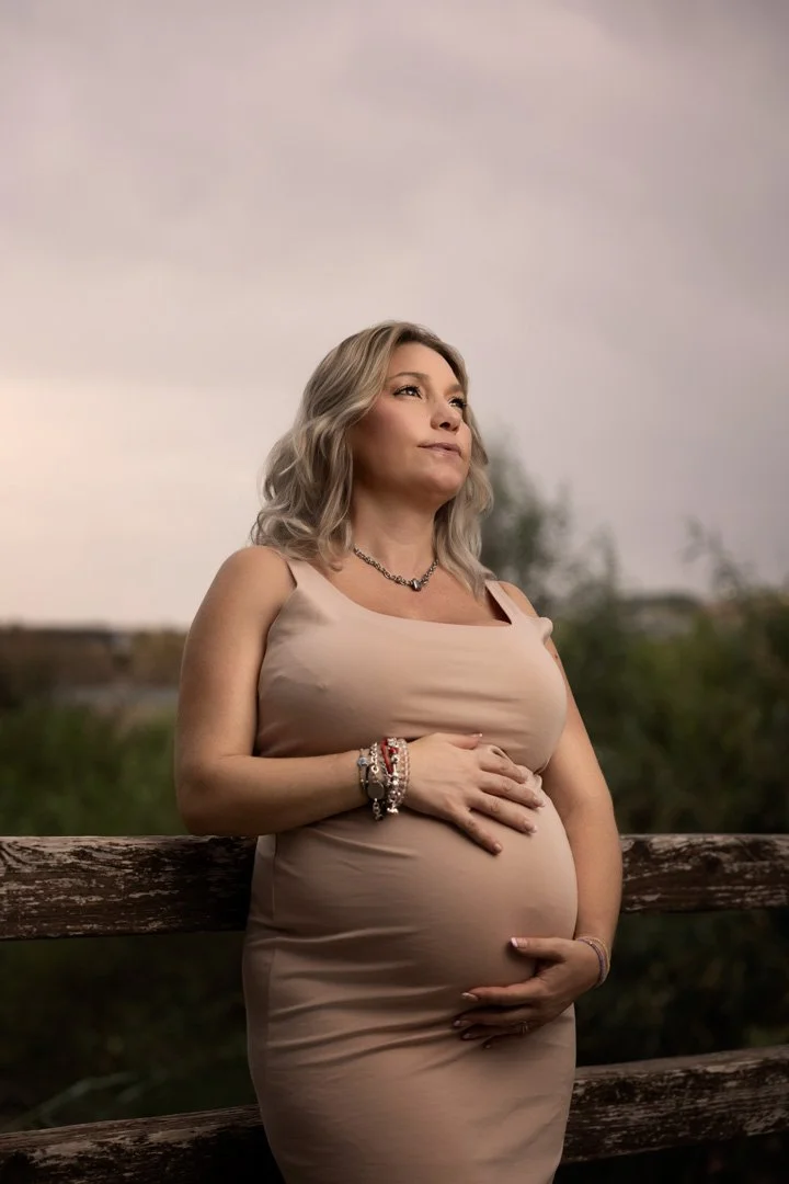 Mujer embarazada con vestido beige, vistas al horizonte, sosteniendo su vientre con ambas manos, en un entorno natural al atardecer.