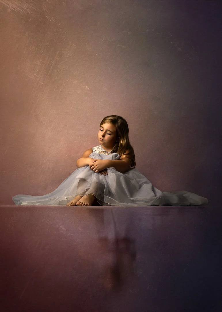 Niña pequeña sentada en el suelo con vestido de color claro, con expresión pensativa o triste, en un entorno con fondo marrón y superficie reflectante.