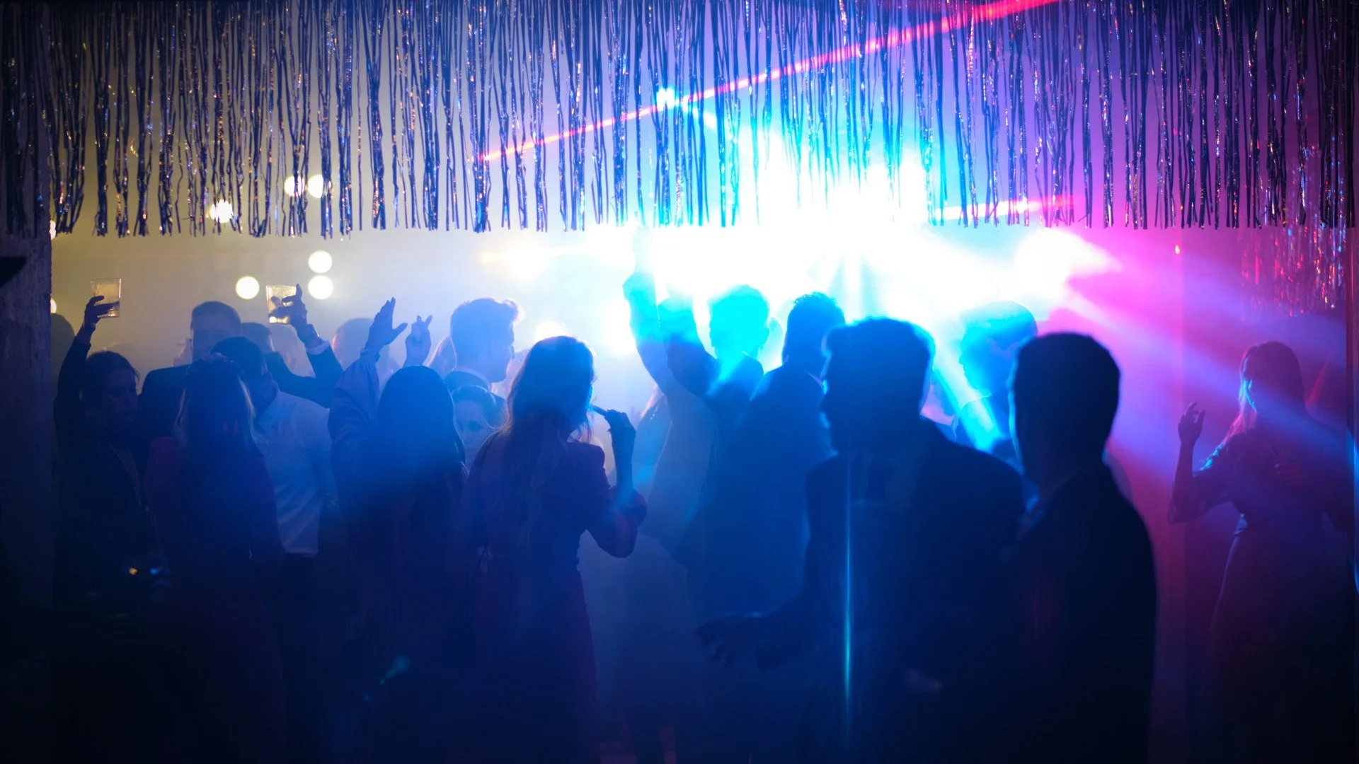 Personas bailando en una fiesta con luces de colores y decoración con tiras de papel doradas en el techo.