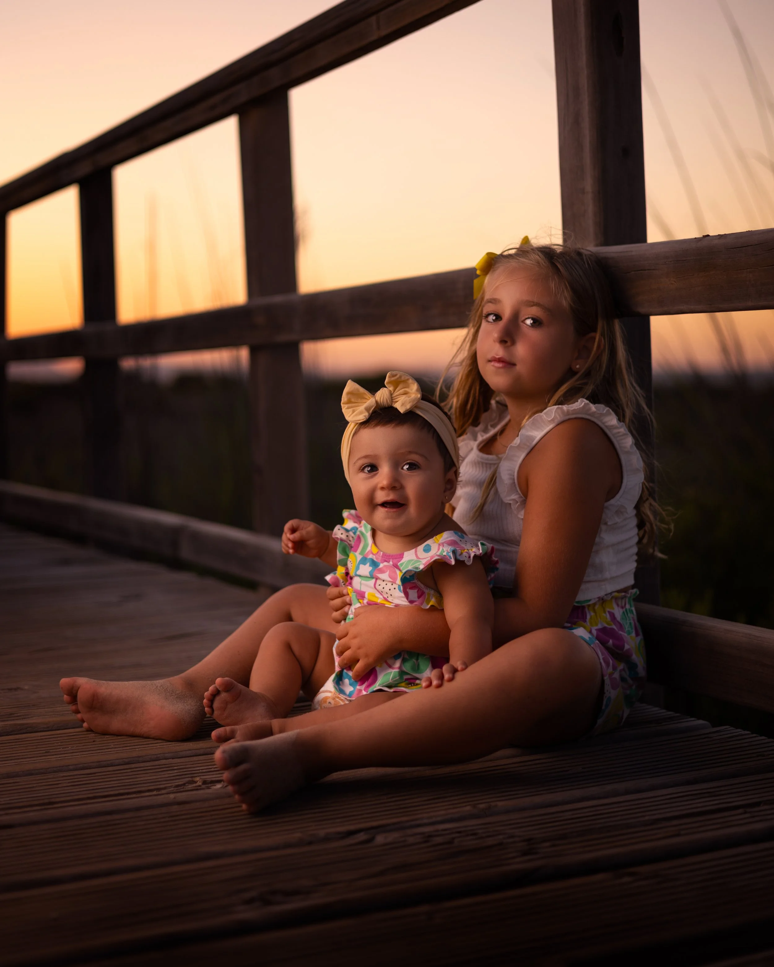 Dos niñas adultas y una bebé sentadas en un muelle de madera al atardecer, la bebé sonríe y la niña mayor observa con expresión serena.