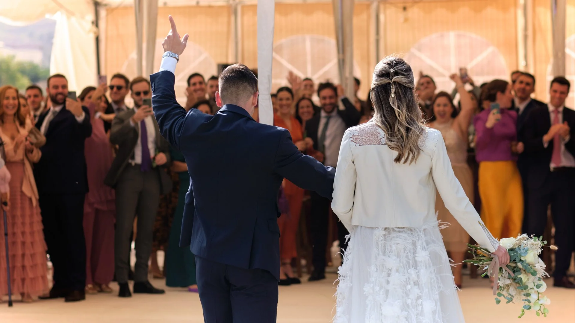 Pareja de recién casados en su boda, el novio con traje azul y la novia con vestido blanco, ella sostiene un ramo de flores y él la acaricia en la espalda, mientras la gente los aplaude y toma fotos.