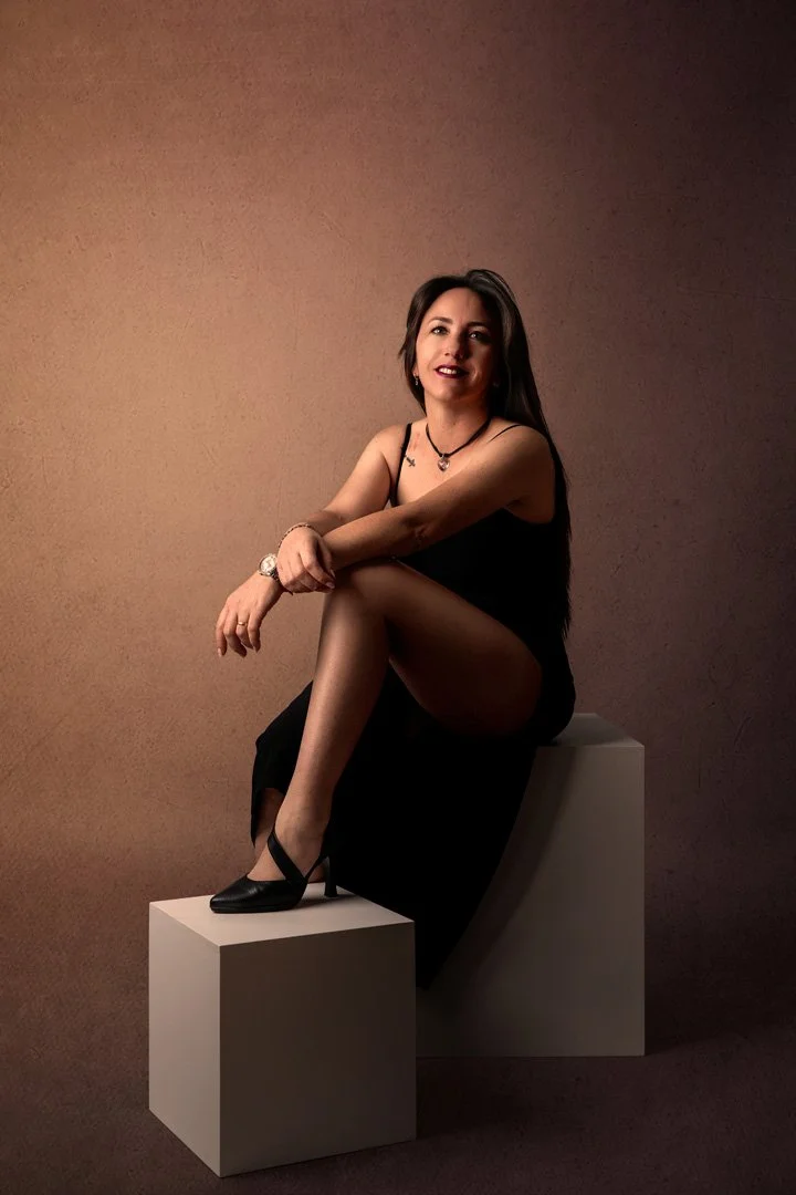 Mujer con vestido negro sentada sobre bloques blancos, posando en estudio con fondo marrón.