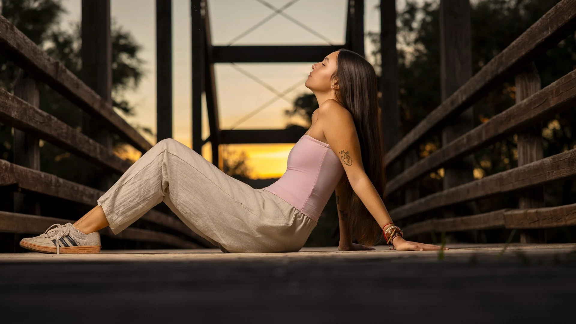 Mujer joven sentada en un puente de madera durante el atardecer, con los ojos cerrados y rostro hacia arriba.
