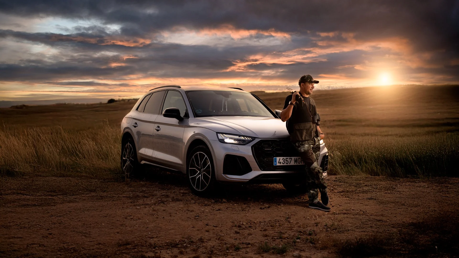 Un hombre con ropa de camuflaje, mochilero y con binoculares, apoyado en un auto SUV de color gris en un campo durante la puesta del sol