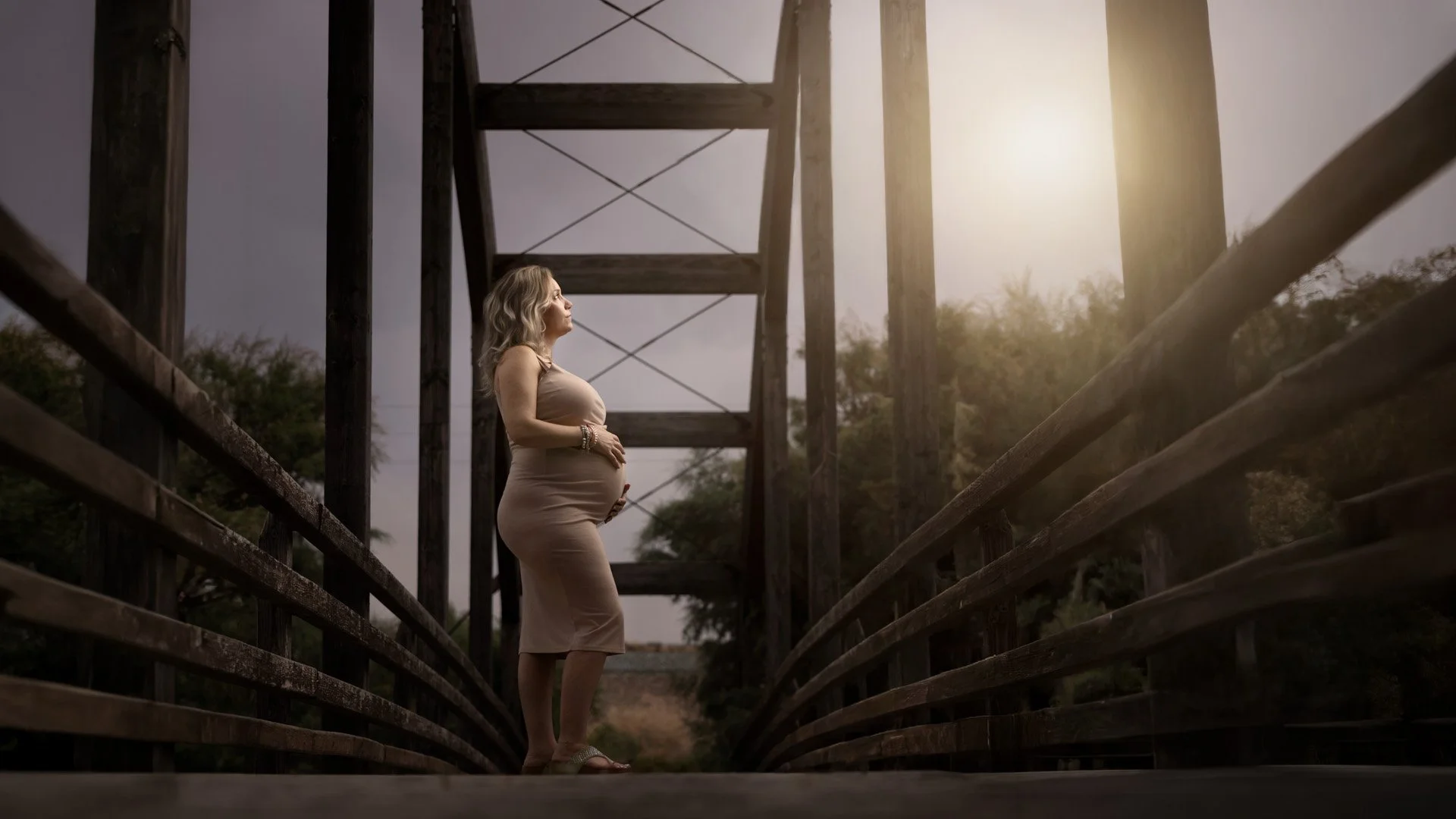 Mujer embarazada de pie en un puente de madera, iluminada por la luz del sol, con árboles en el fondo.