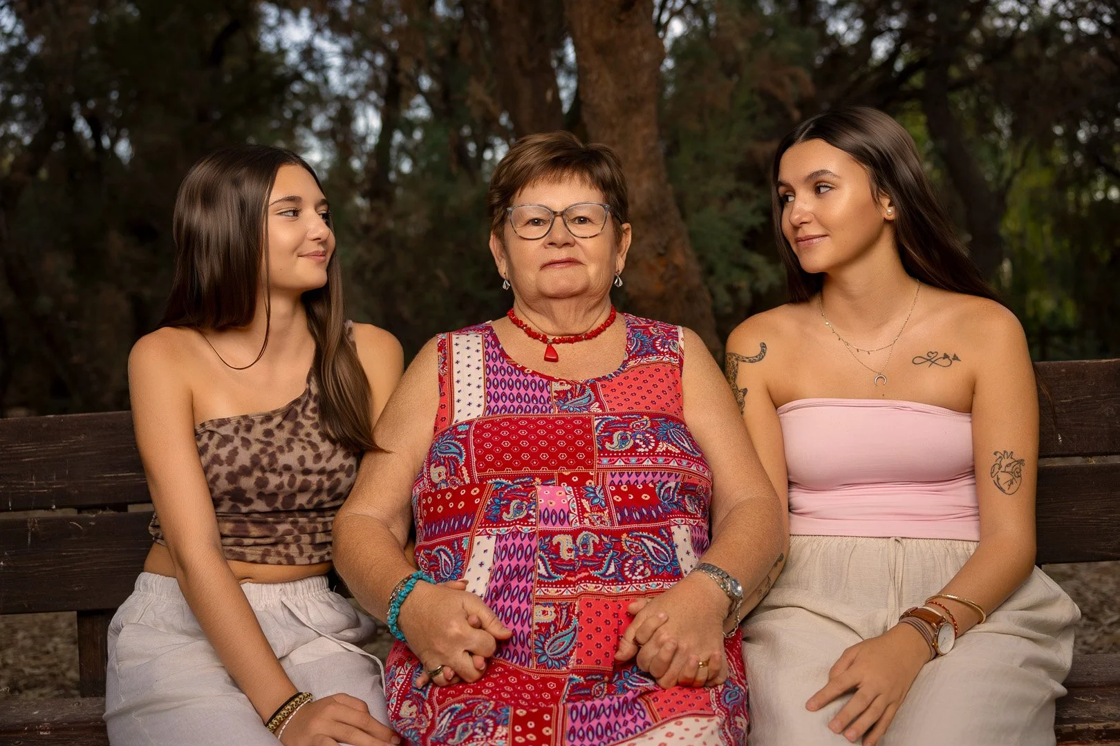 Tres mujeres adultas sentadas en un banco de parque, la mujer del centro es de edad avanzada con vestido colorido, acompañada de dos jóvenes con ropa casual, en un entorno natural con árboles.