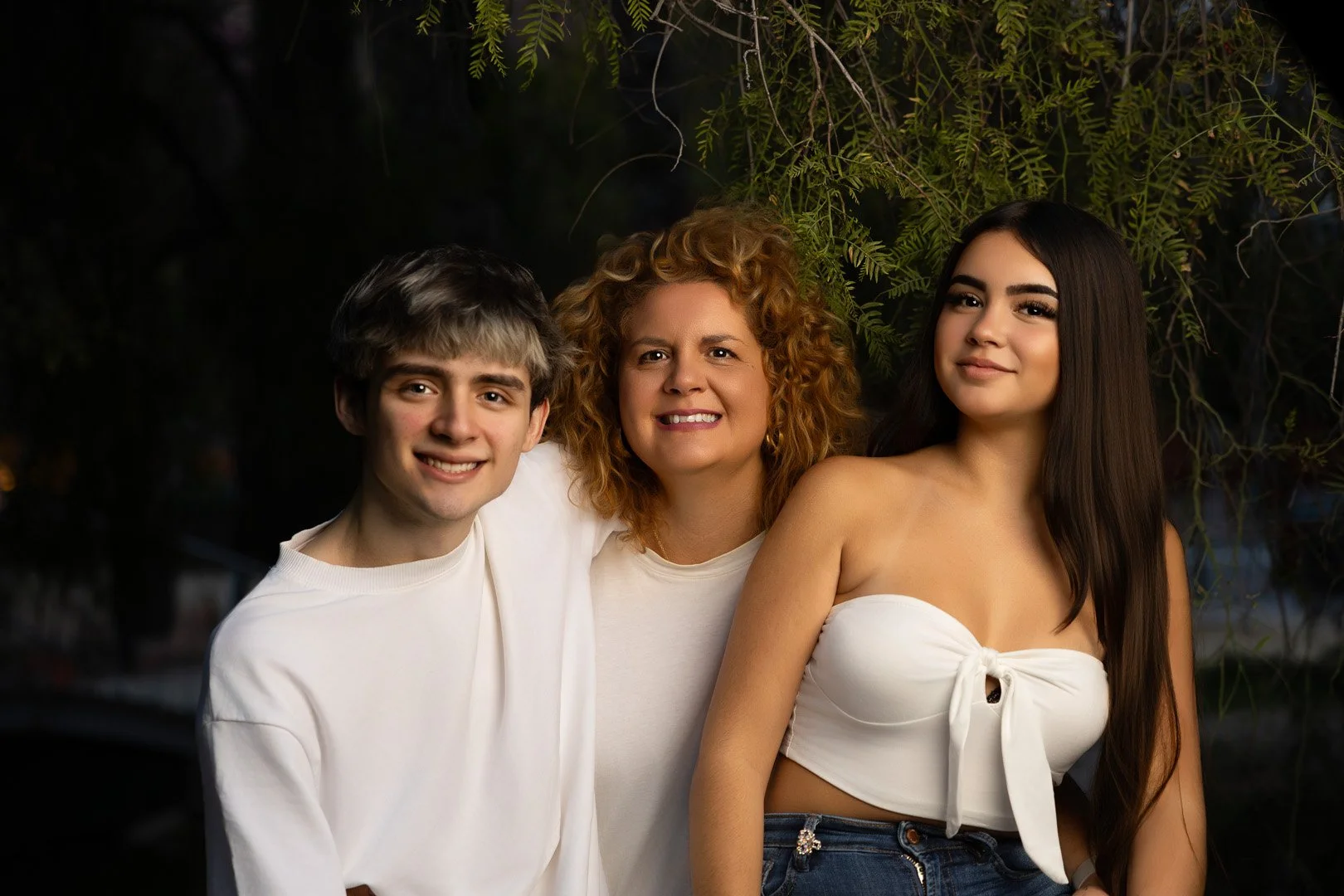 Tres personas jóvenes, un hombre, una mujer con cabello rizado y una mujer con cabello largo y liso, posando juntas al aire libre en la noche, con ramas verdes en el fondo.