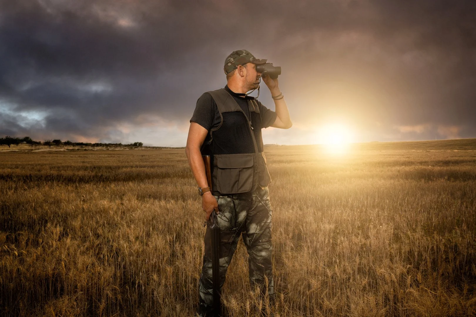 Un hombre en un campo de trigo observa usando binoculares durante el atardecer o amanecer, lleva ropa de caza y mochila.
