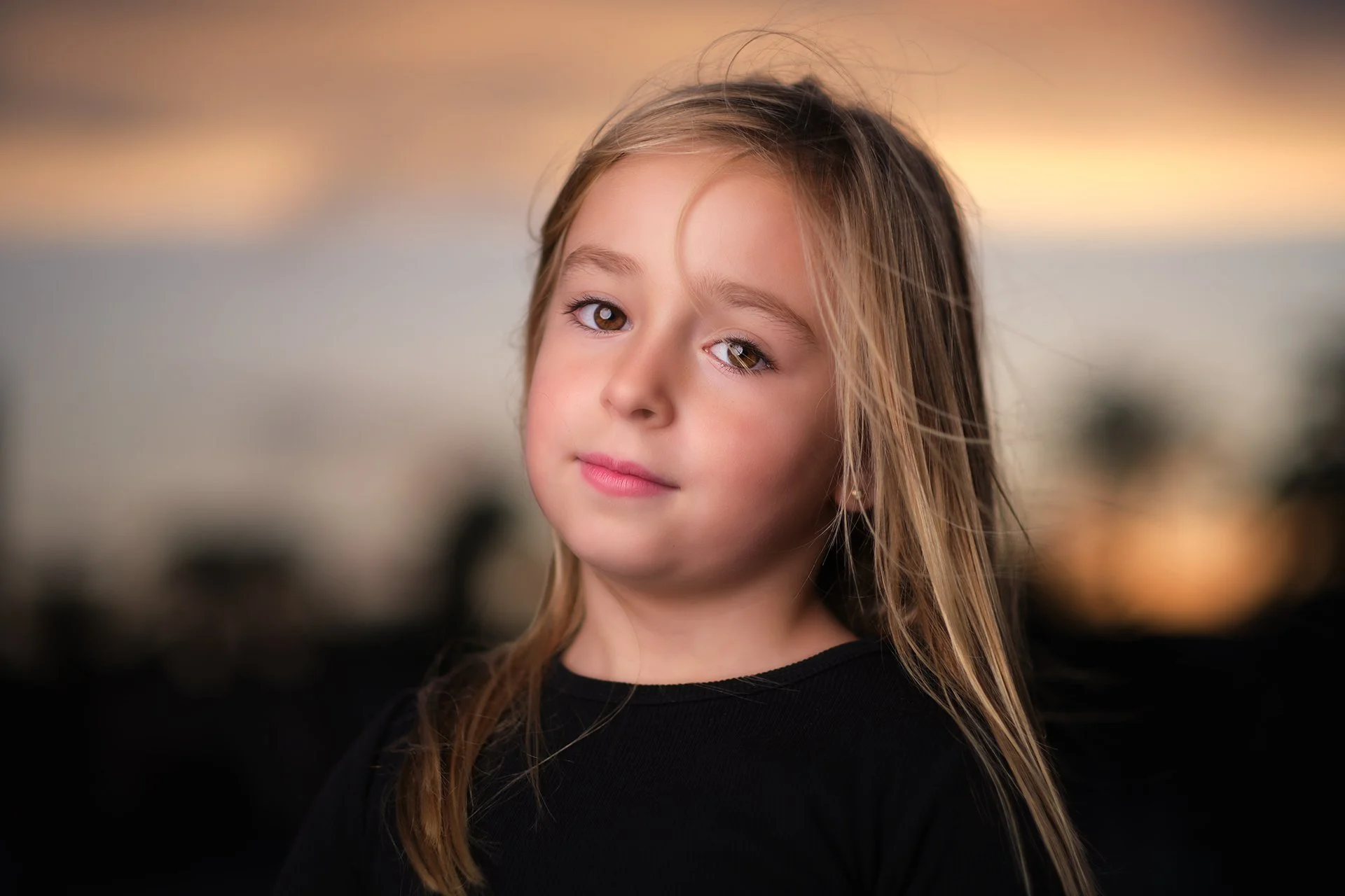 Retrato de una niña con cabello rubio, ojos claros, con expresión tranquila, de fondo difuminado con tonos beige y anaranjados, al atardecer.