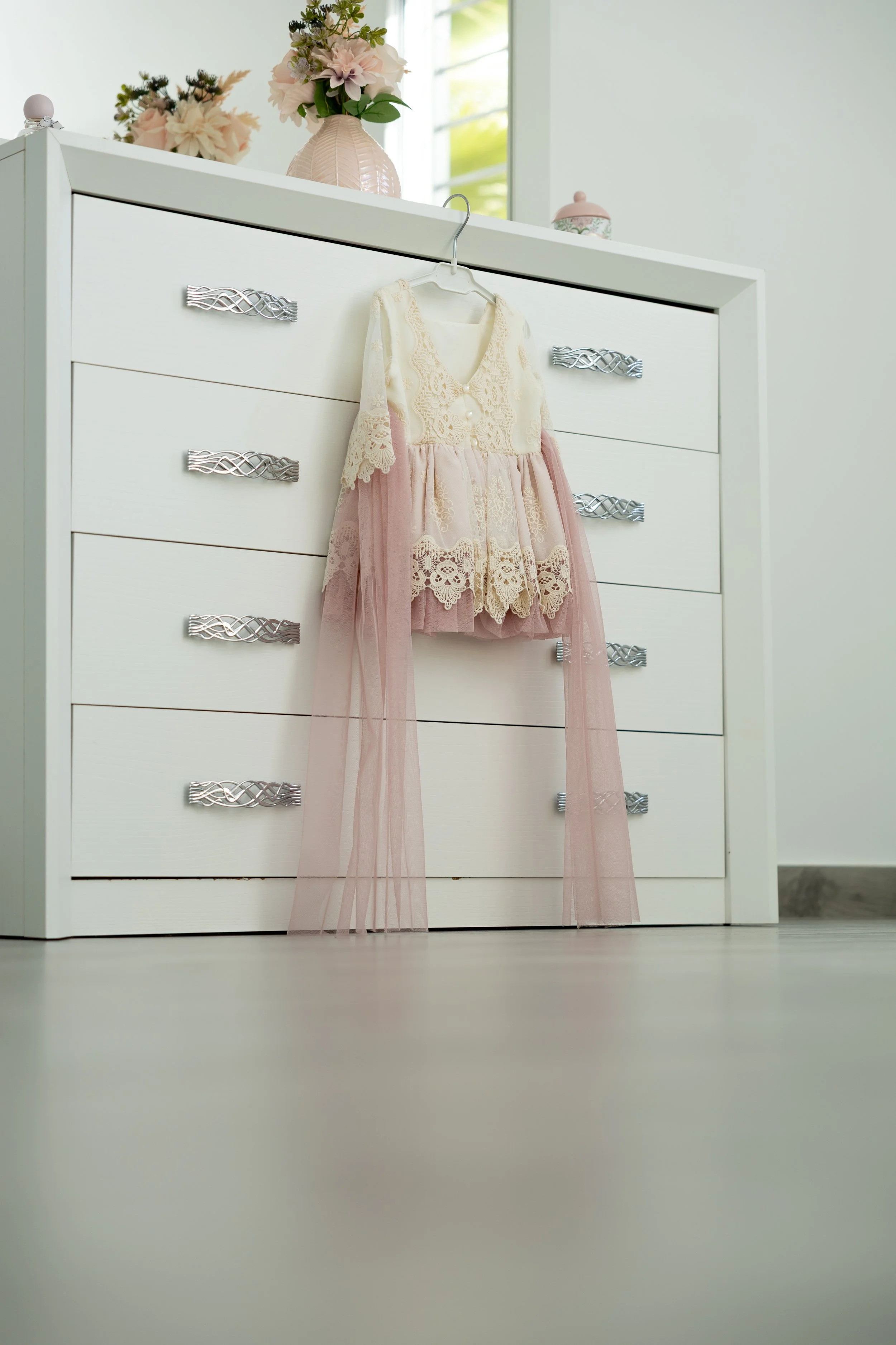 Ropa de bebé con encaje y tul rosa colgando en una percha sobre un mueble blanco con cajones.