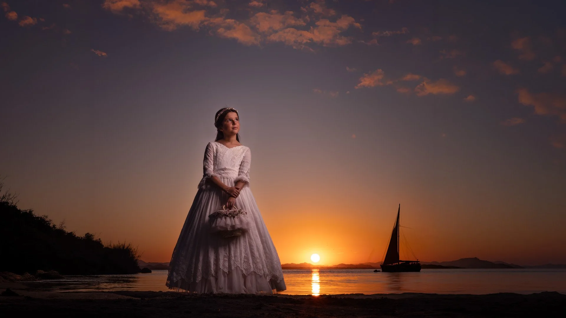 Una niña vestida con un vestido blanco de encaje y una diadema en el cabello, de pie en la orilla del mar durante el atardecer, con un barco a vela en el agua y montañas a lo lejos.