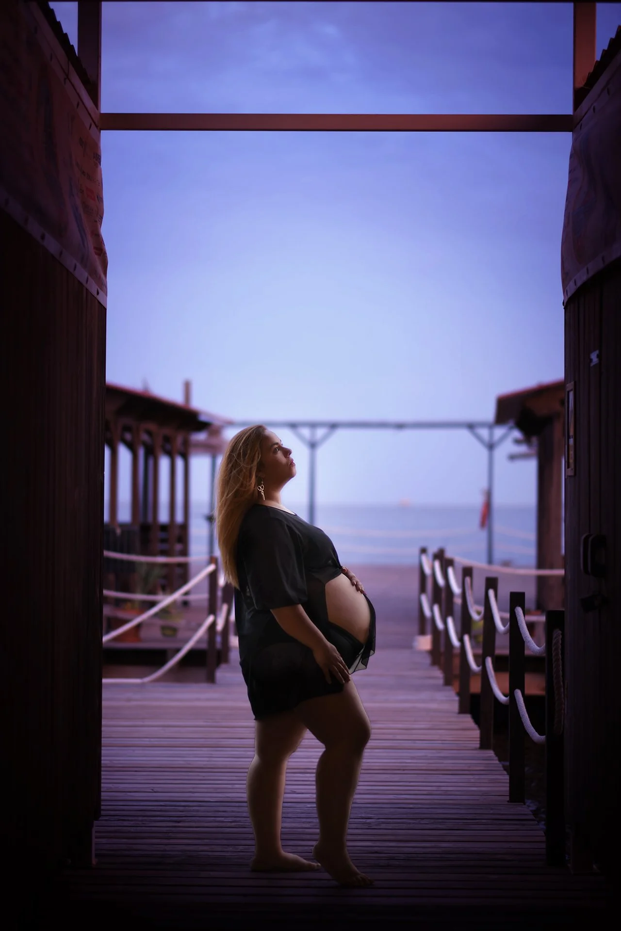 Una mujer embarazada de pie en una plataforma de madera en la playa durante el atardecer, con el mar de fondo y el cielo con tonos azules y violetas.
