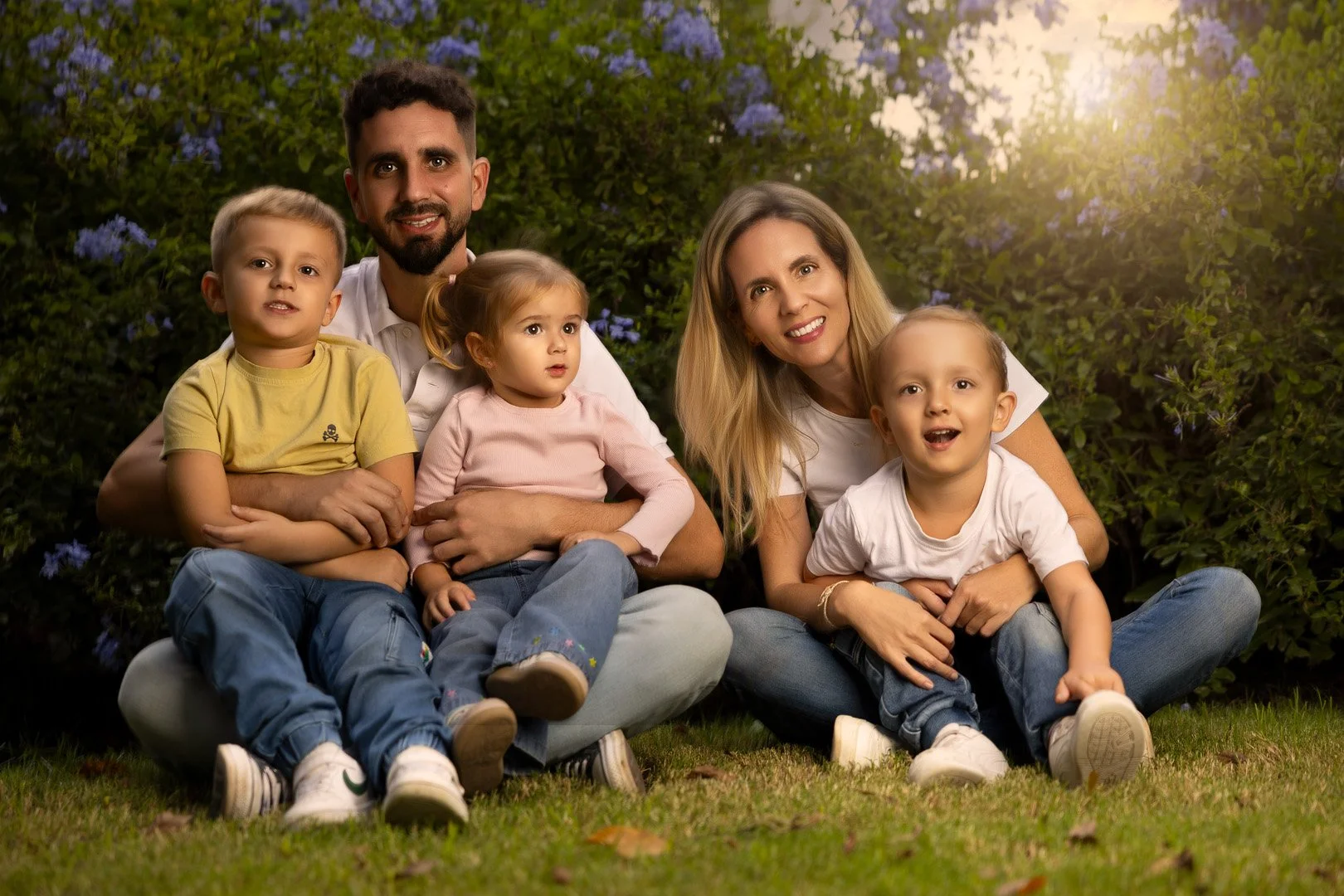Familia sentada en el césped, rodeada de plantas con flores, con luz cálida de puesta de sol al fondo.
