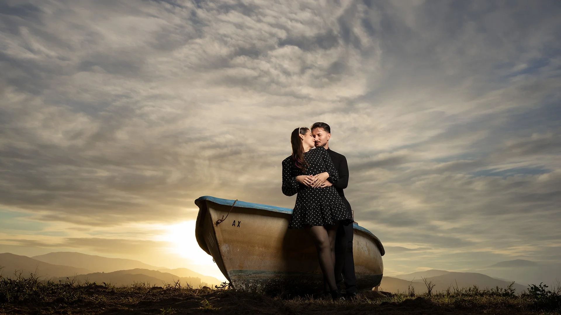 Una pareja abrazada en un bote abandonado en un campo con montañas y un cielo nublado al atardecer.