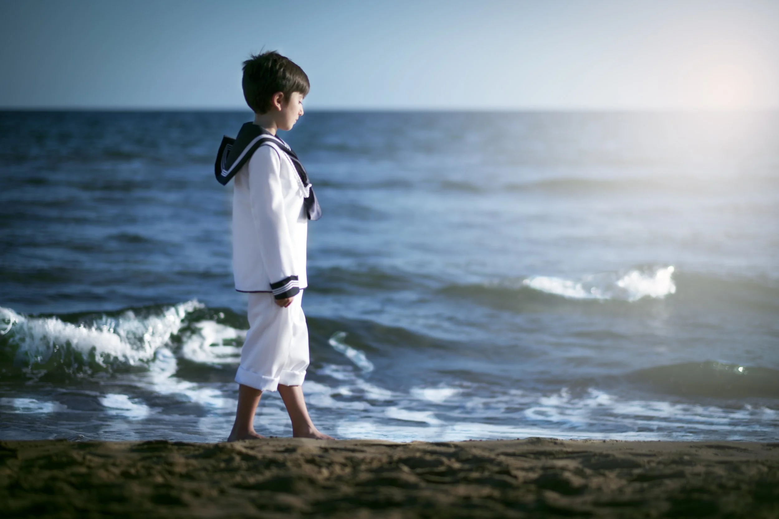 Un niño vestido con uniforme escolar en la playa, mirando hacia el mar en un atardecer