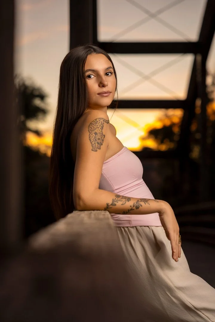 Retrato de una mujer joven con tatuajes, usando un top rosa y una falda beige, en un entorno al atardecer con estructuras metálicas y árboles al fondo.