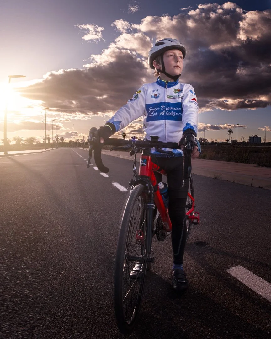 Joven en bicicleta con casco y ropa deportiva en una carretera al atardecer con nubes oscuras y palmeras en el fondo.