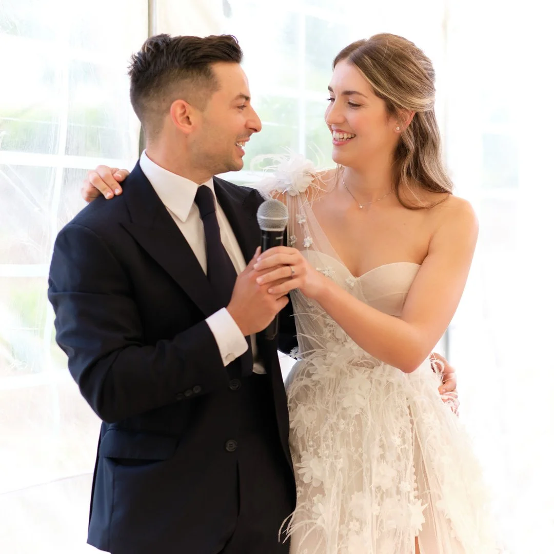 Una pareja de recién casados riendo y abrazados en una boda, ella con vestido de novia y él con traje formal, sosteniendo un micrófono.
