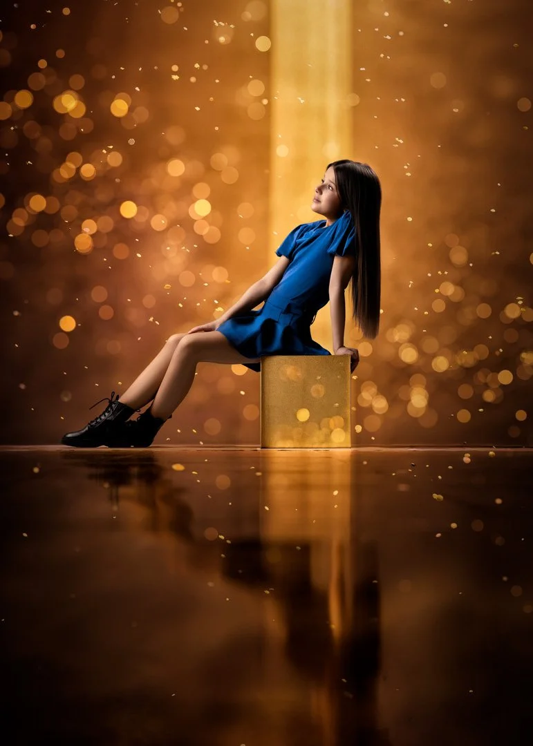 Una niña con vestido azul y botas negras sentado en un bloque dorado, mirando hacia arriba, fondo dorado con efectos de bokeh y reflejo en el suelo.