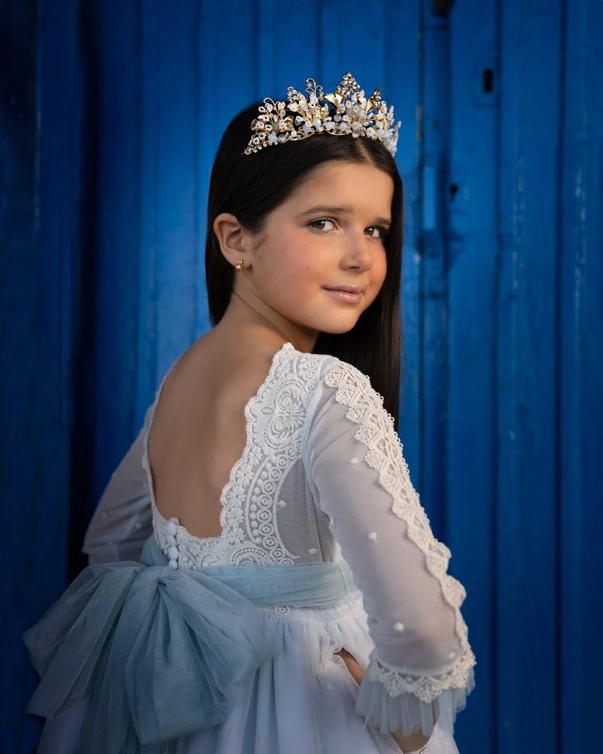 Jovencita con vestido blanco, diadema de perlas y cabello largo, frente a fondo de madera azul.