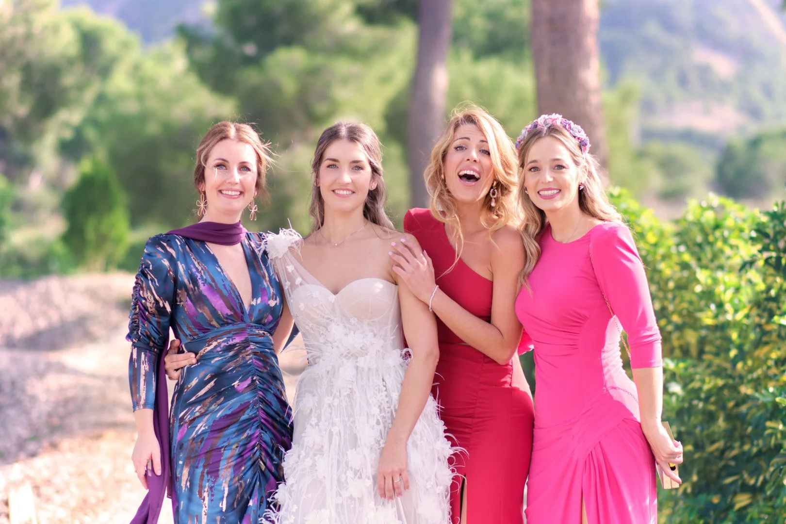 Cuatro mujeres en vestidos de fiesta sonrientes en un entorno al aire libre con árboles.