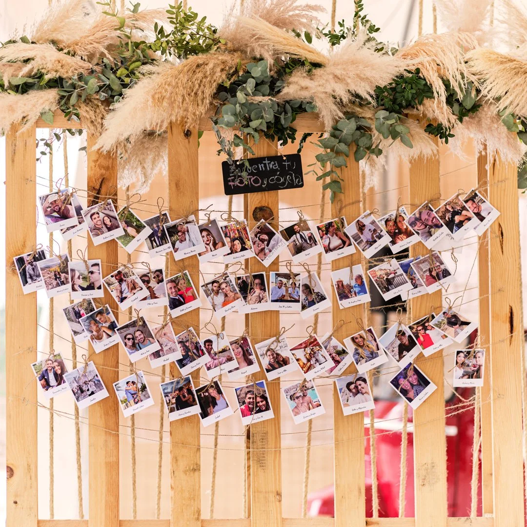Decoración de boda con fotos de los invitados colgadas en cuerdas en un marco de madera adornado con follaje y plumas.