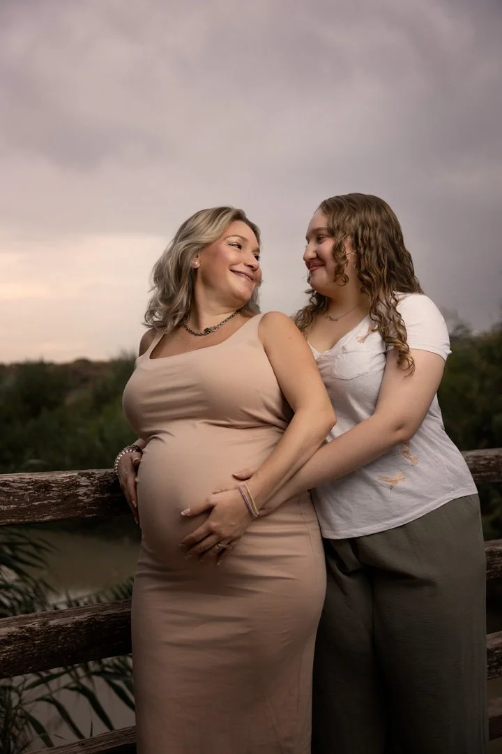 Dos mujeres sonriendo y mirándose con ternura, una embarazada y la otra probablemente su hija, en un paisaje exterior con cielo nublado y vegetación al fondo.