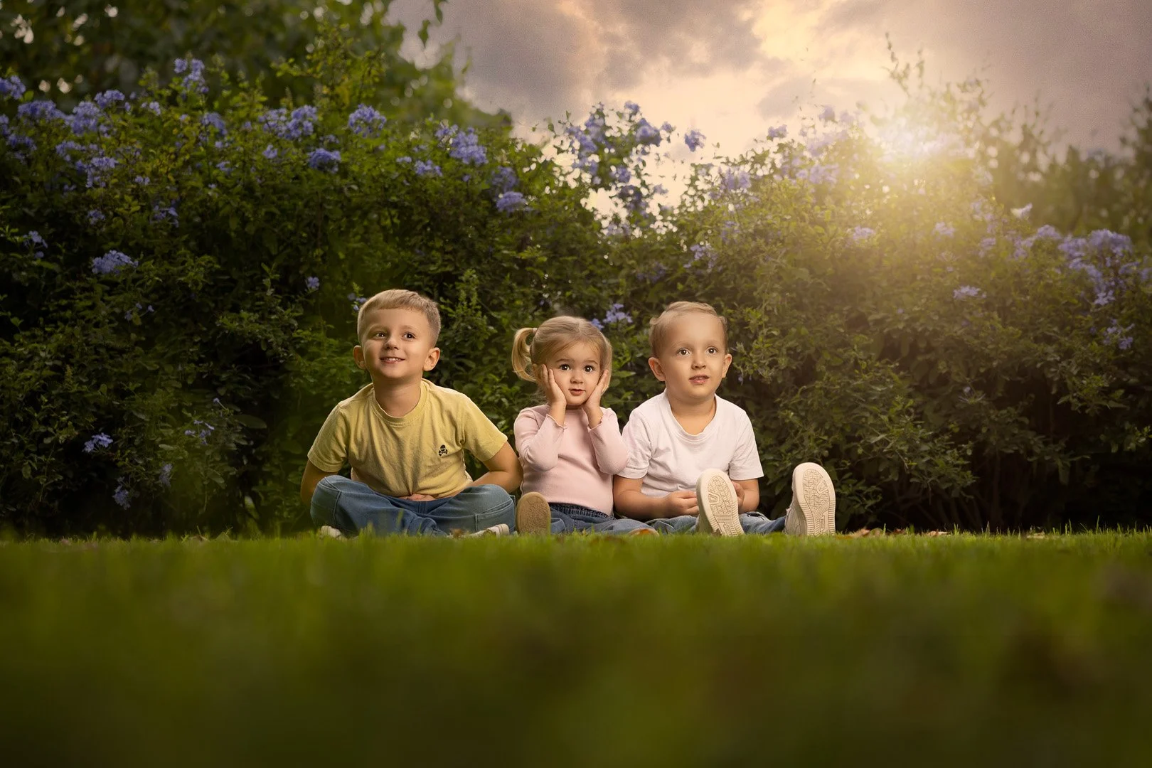 Tres niños sentados en la hierba en un día soleado, con arbustos con flores moradas y un cielo despejado con nubes, disfrutando de la naturaleza.