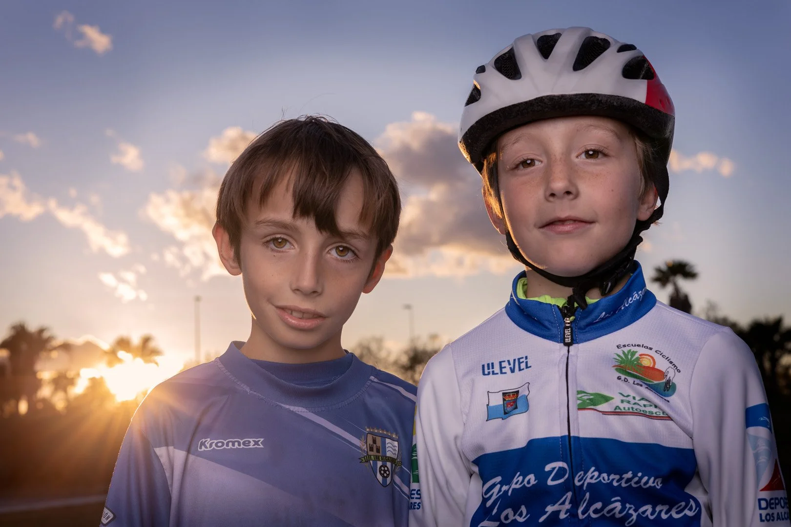 Dos niños con ropa de ciclismo posando al atardecer, uno con casco y ambos con expresiones tranquilas, en un entorno con árboles y cielo con nubes.