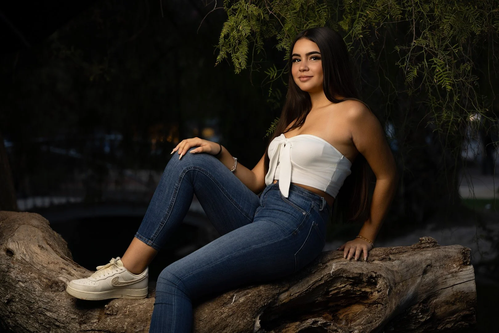 Joven mujer con blusa blanca y jeans sentada sobre tronco de árbol en escena de noche en naturaleza