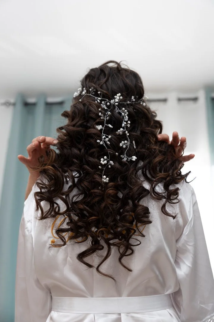 Cabello rizado con adornos florales y perlas visto desde atrás, con una bata blanca y cortinas azules en el fondo.