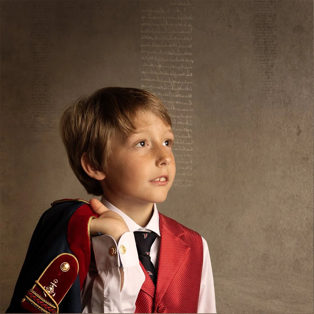 Un niño con uniforme escolar sostenido en su hombro un saco de color negro con rojo y detalles dorados, mira hacia arriba en actitud pensativa, con una pared de fondo que tiene escritos antiguos en color sepia.