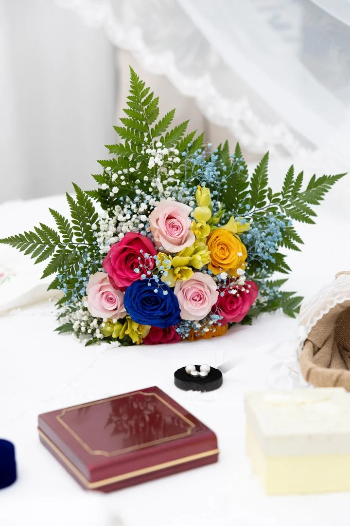 Ramo de flores con rosas de varios colores, helechos y pequeñas flores blancas y azules, sobre una mesa blanca.