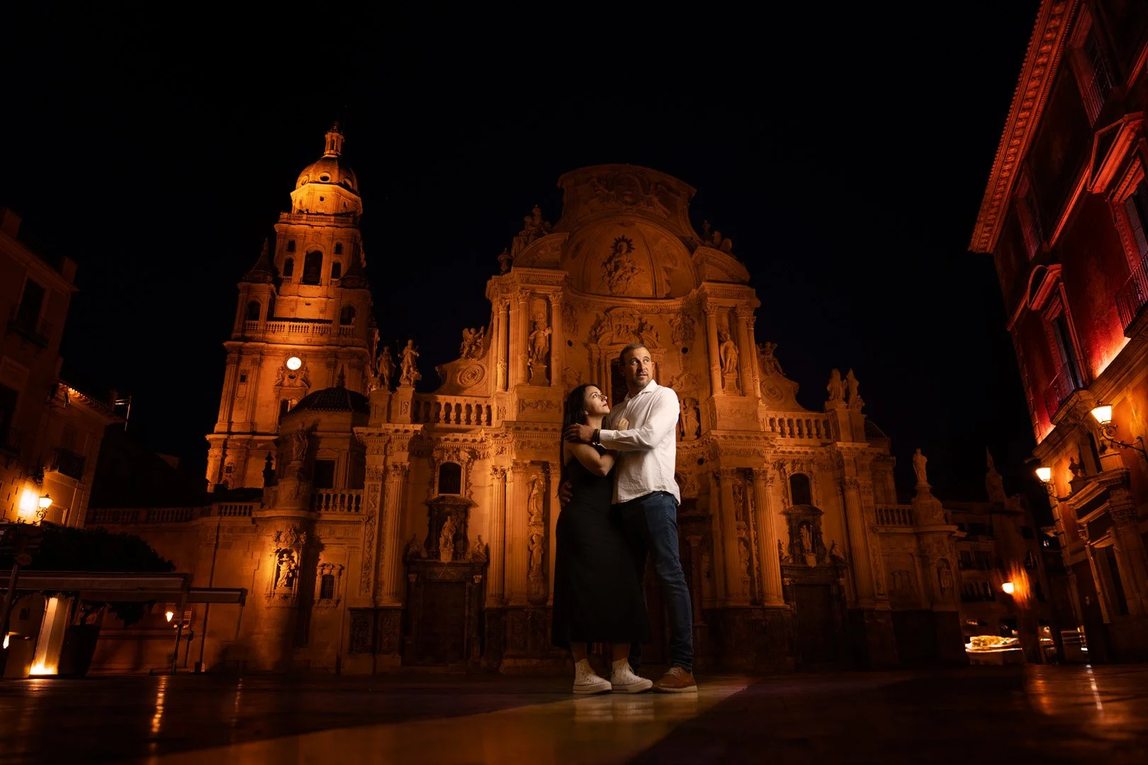Una pareja abrazada en una plaza iluminada por la noche, con una catedral antigua de fondo.