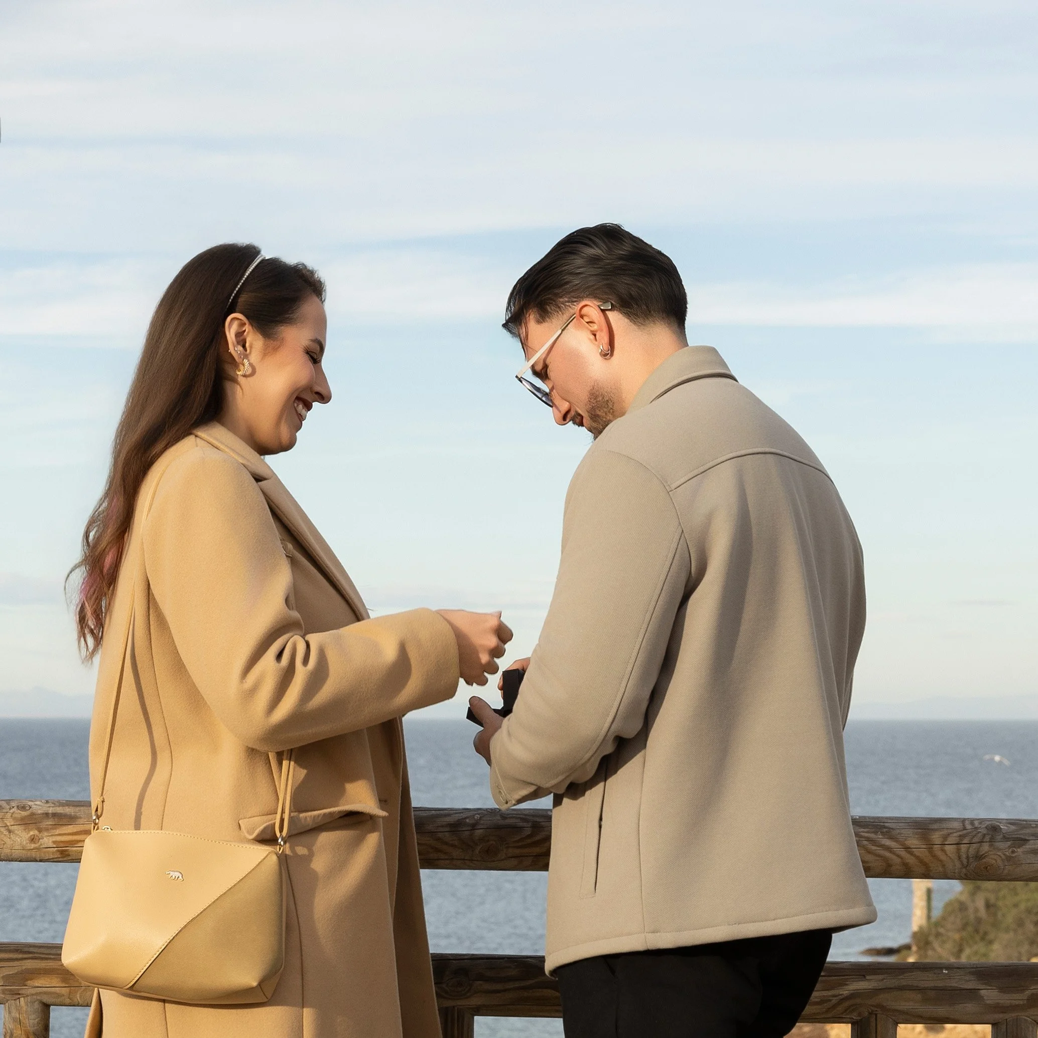 Una pareja en un muelle junto al océano, intercambiando el anillo de compromiso, ambos sonriendo y vestidos con ropa beige.