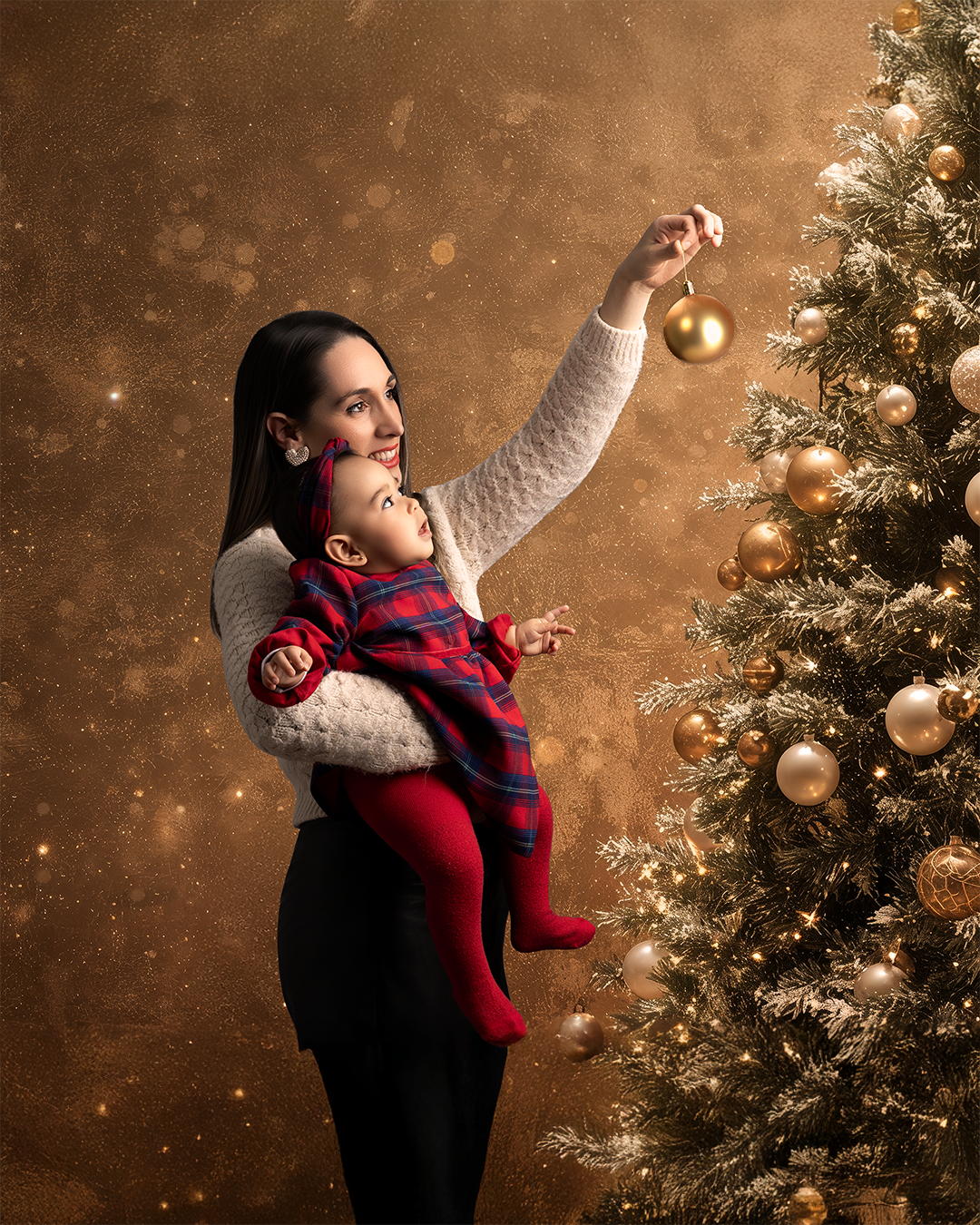 Una mujer joven con un niño pequeño decorando un árbol de Navidad con esferas doradas en un fondo dorado con destellos.
