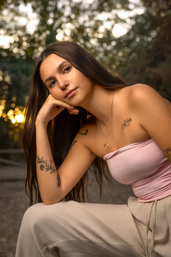 Jovencita con tatuajes en brazos y pecho, usando top rosa claro y pantalones beige, posando en naturaleza al atardecer.