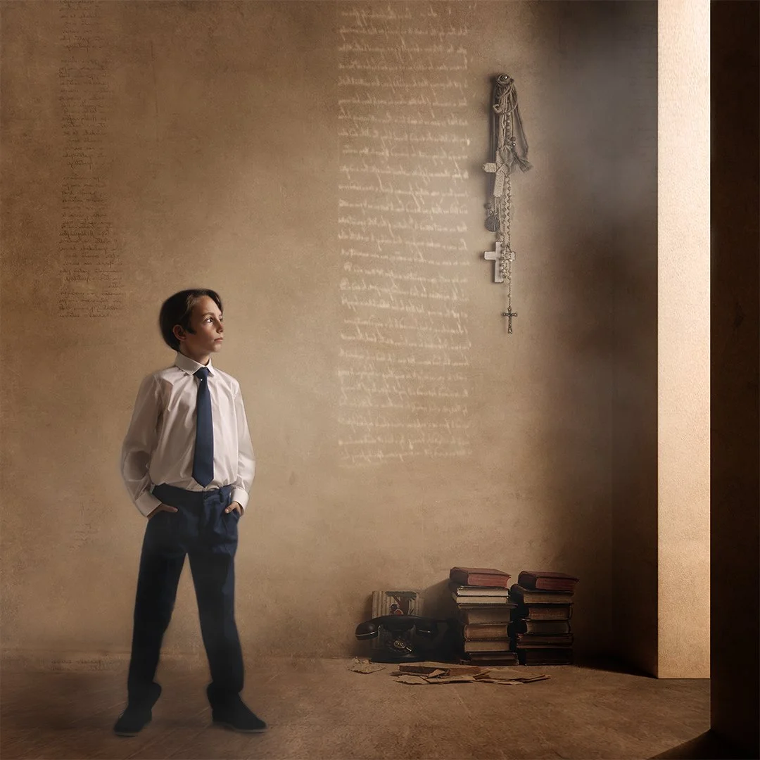 Un niño con camisa blanca, corbata azul y pantalones oscuros, en una habitación con paredes de color beige, mirando hacia la izquierda, con libros apilados en el suelo y cadenas de rosarios y crucifijos colgados en la pared.