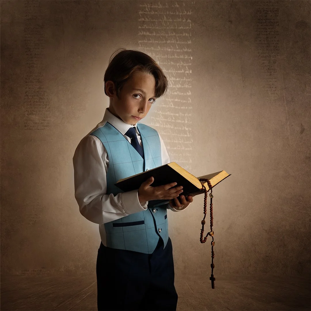 Joven niño sosteniendo un libro abierto, vestido con camisa blanca, chaleco azul y corbata, con un rosario en la mano, en un fondo con texto decorativo.