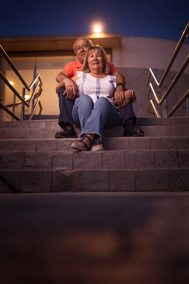Una pareja de adultos mayores sentados en las escaleras durante la noche, sosteniendo las manos y sonriendo.