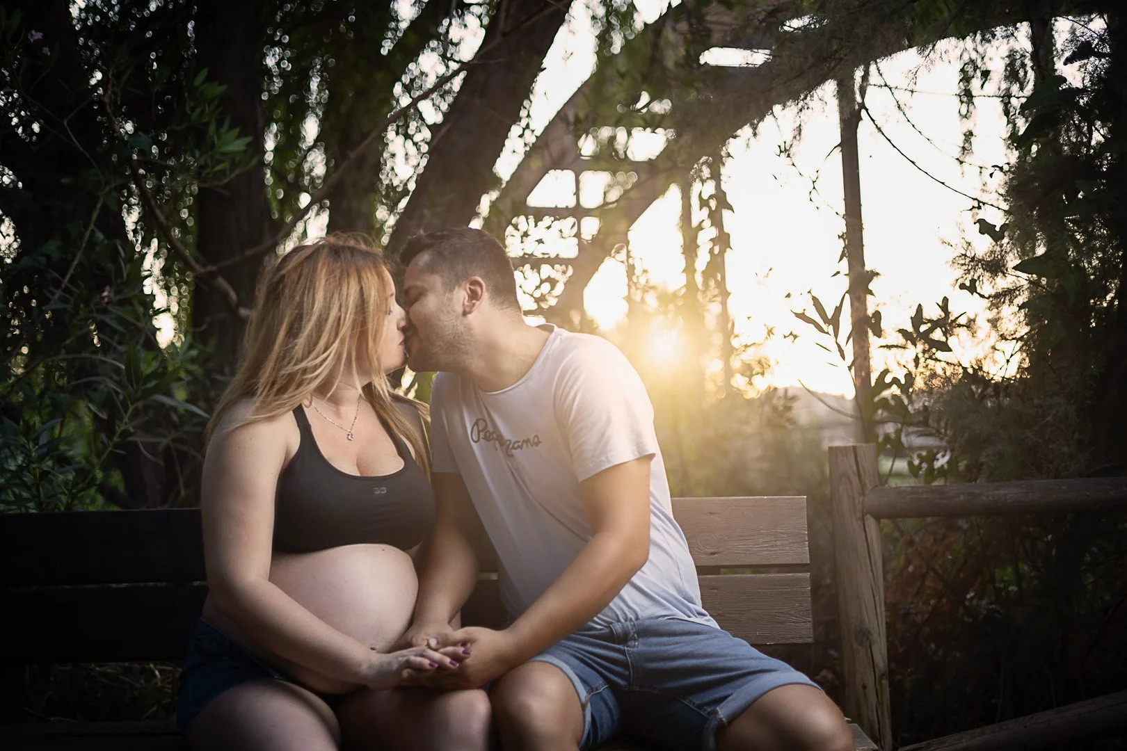 Una pareja en un banco de madera compartiendo un beso, la mujer embarazada, al atardecer en un entorno natural con árboles y sombra.