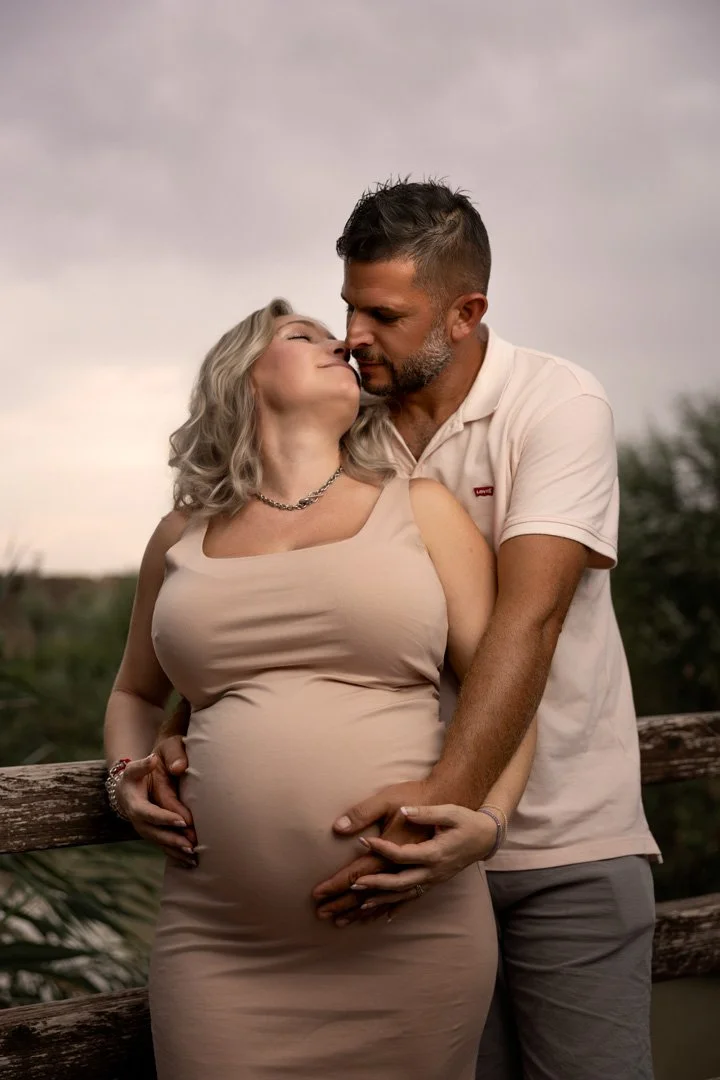 Una pareja abrazada, con una mujer embarazada, en un entorno al aire libre, en un puente de madera, en un día nublado, compartiendo un momento íntimo y cariñoso.