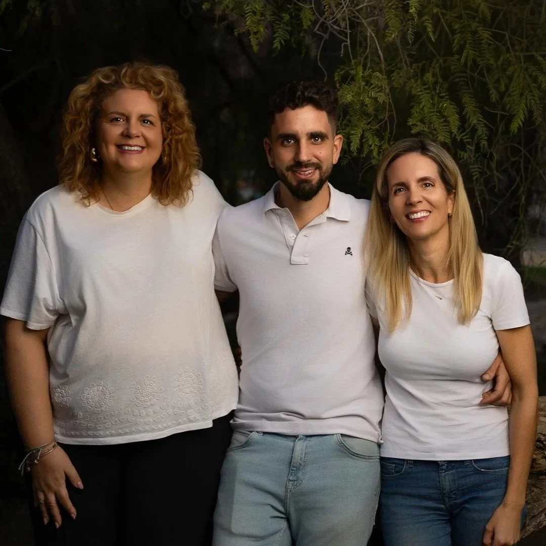 Tres personas (dos mujeres y un hombre) con ropa blanca, sonriendo y de pie al aire libre con árboles en el fondo.