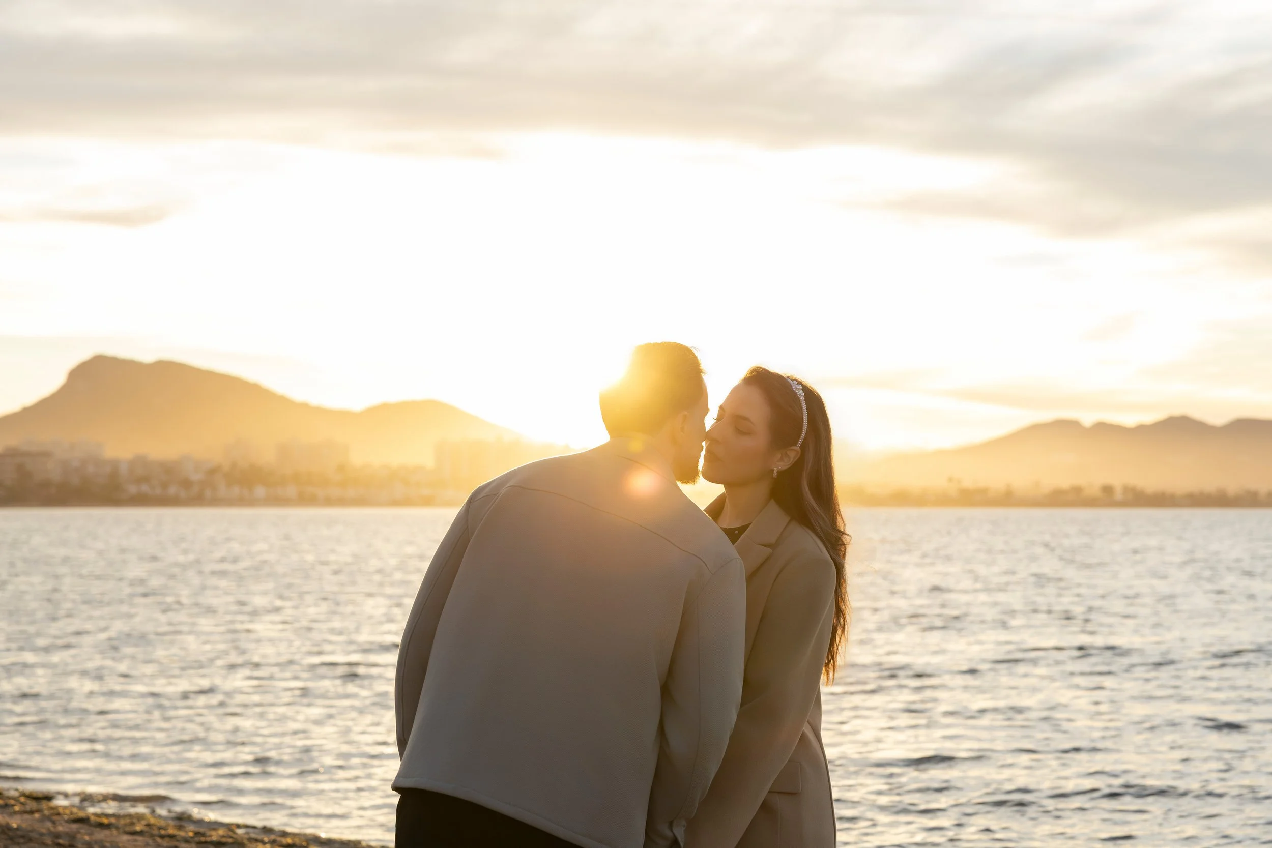 Una pareja está a punto de besarse en la orilla de un lago o mar, con montañas de fondo, durante el atardecer, con el sol brillante y un cielo nublado.