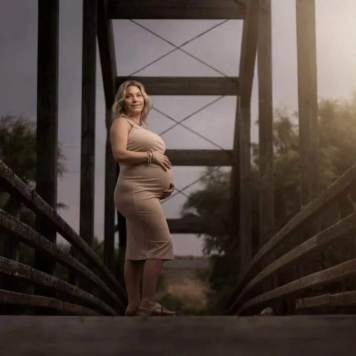 Mujer embarazada de cabello rubio y largo, vestida con un vestido sin mangas beige, de pie en un puente de madera, con una estructura de torres de fondo, en un entorno natural con árboles y luz cálida.