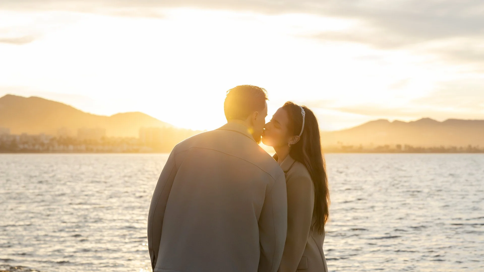 Pareja enamorada besándose al atardecer frente a un lago con montañas al fondo.