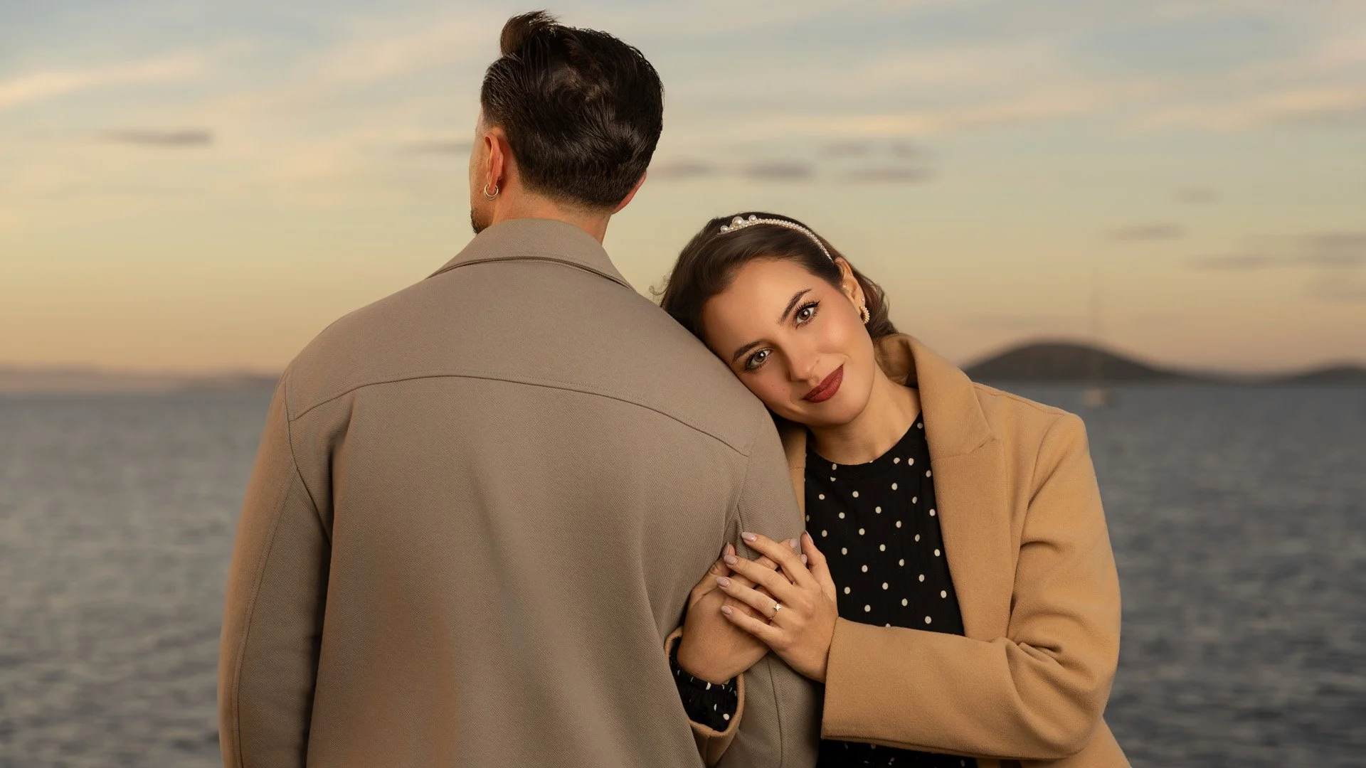 Una mujer joven con cabello rizado y vestido negro con puntos blancos, sonriendo, apoya su cabeza en el hombro de un hombre con cabello oscuro y rizado, usando un abrigo beige, en un entorno junto a un cuerpo de agua durante el atardecer.